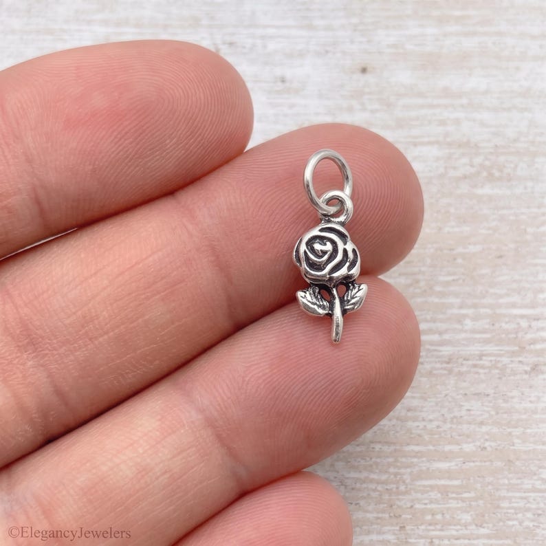 May include: A small silver-toned metal charm shaped like a rose flower with a single stem and two leaves. The charm features a circular jump ring at the top for attachment. The design has oxidized detailing to highlight the petals and stem structure.