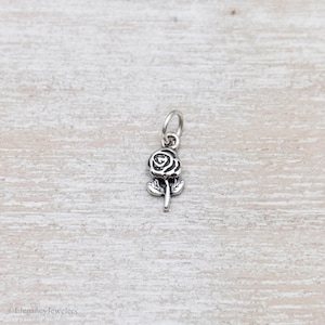May include: A small silver-toned metal charm shaped like a rose flower with a stem and two leaves. The charm features oxidized detailing in the recessed areas to define the petals and foliage, and it includes a circular jump ring at the top.