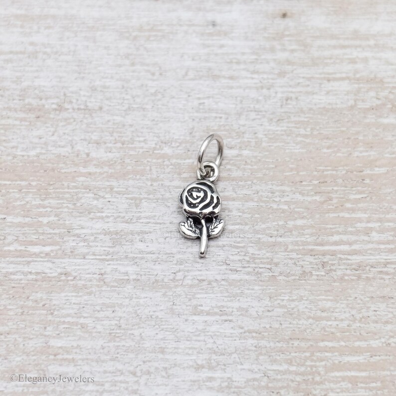 May include: A small silver-toned metal charm shaped like a rose flower with a stem and two leaves. The charm features oxidized detailing in the recessed areas to define the petals and foliage, and it includes a circular jump ring at the top.