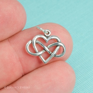 May include: Silver charm in the shape of a heart with an infinity knot design. The charm is shown on a finger.