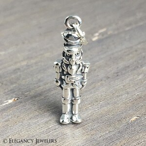 May include: A silver charm depicting a toy soldier nutcracker. The charm features intricate detailing of a hat, uniform, and boots. It hangs from a small loop at the top. The background is a wooden surface.