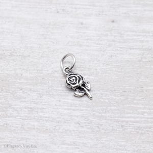 May include: A small, silver-toned metal charm shaped like a rose flower with a single leaf attached to the stem. The charm features an attached circular jump ring at the top for hanging. The design has darkened, recessed details to highlight the rose petals.