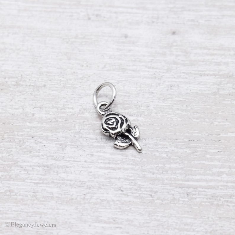 May include: A small, silver-toned metal charm shaped like a rose flower with a single leaf attached to the stem. The charm features an attached circular jump ring at the top for hanging. The design has darkened, recessed details to highlight the rose petals.