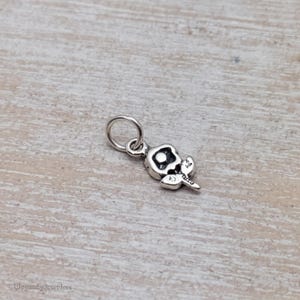 May include: A small silver-toned metal charm shaped like a stylized flower with a circular center and a single leaf extending from the base. The charm features an attached jump ring at the top for hanging, with oxidized detailing to define the floral shape.