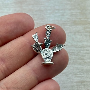 Puede incluir: Un charm de cactus plateado con un diseño detallado. El charm presenta una forma de nopal con superficies texturizadas y un pequeño lazo para sujetarlo a un collar o pulsera. El charm se sostiene en una mano.