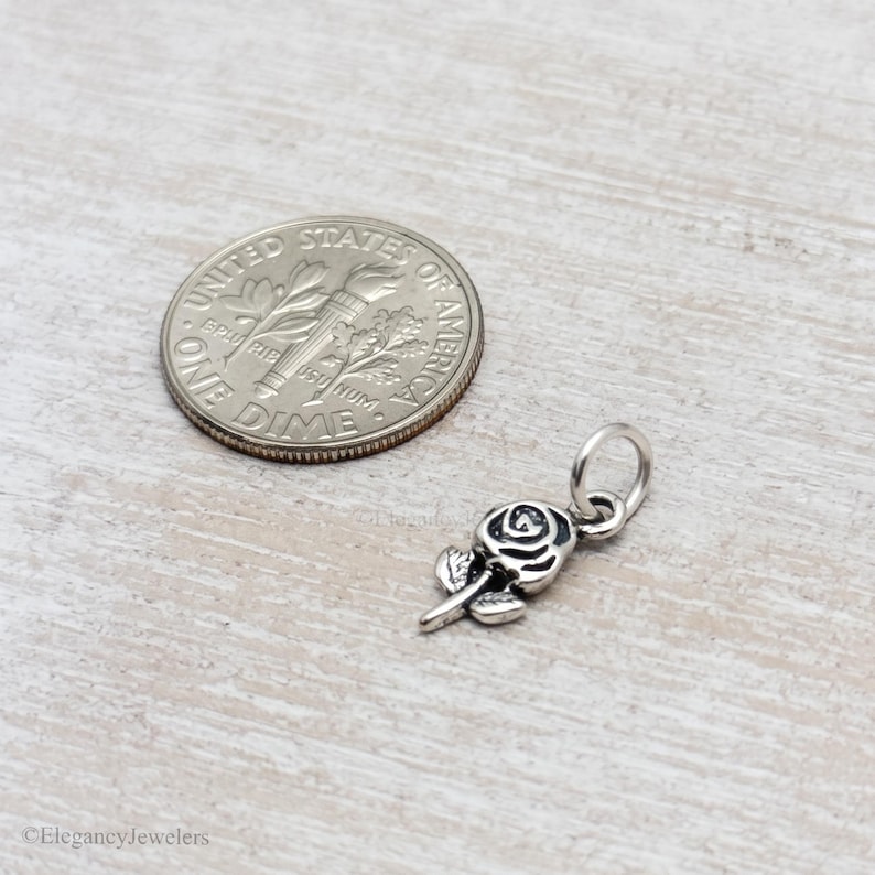 May include: A small silver-toned metal rose charm featuring detailed petal engravings and a circular jump ring at the top. The charm is positioned next to a United States dime for scale, showing its compact size relative to the 0.705-inch coin.
