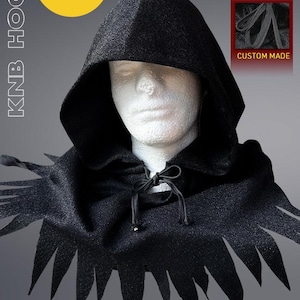 May include: A black KNB hood, prop size, with a pointed hood and decorative, jagged edges. The hood has drawstrings and is custom made. The text "ALL-NEW 2025 VERSION!!" is in a yellow circle. The text "WEARABLE / DISPLAYABLE COLLECTIBLE" is also visible.