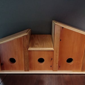 May include: Three wooden birdhouses with slanted roofs and round entry holes. The birdhouses are stacked on top of each other and are painted in a natural wood color.