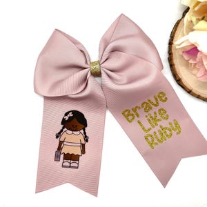 May include: A pink hair bow with a gold glitter design. The bow has a cartoon image of a young girl with brown skin and braids. The text "Brave Like Ruby" is written in gold glitter on the bow.