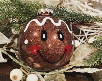 May include: A round, brown glittery Christmas ornament featuring a white icing-style wavy line at the top, black oval eyes with eyelashes, two small red heart-shaped cheeks, and a thin black curved line forming a smiling mouth.