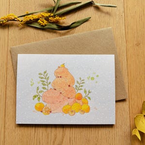 May include: A greeting card with a whimsical illustration of a stack of orange-colored creatures, surrounded by oranges and green foliage. The card is on a wooden surface with a brown envelope and decorative yellow flowers.