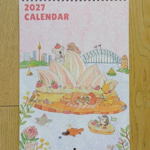 May include: A 2027 calendar featuring a watercolour illustration of the Sydney Opera House styled as a dessert with strawberries and cream. Various animals including a koala, kangaroo, and platypus are depicted around the structure.