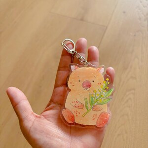 May include: A clear acrylic keychain featuring a watercolor illustration of a wombat holding yellow flowers. The wombat is orange with red accents on its feet and nose. The keychain has a silver metal ring.