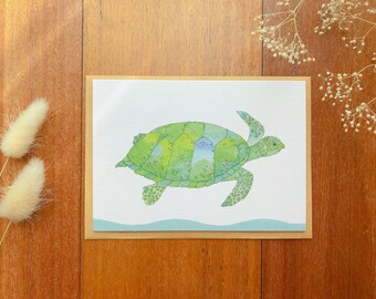 May include: A watercolor greeting card featuring a green sea turtle illustration. The turtle has a detailed shell with shades of green and blue. The card is on a white background with a light blue wave design, set in a brown envelope.