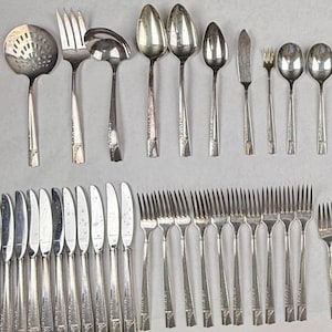 May include: A collection of vintage silver-colored flatware, including spoons, forks, knives, and serving utensils. The arrangement showcases various sizes and styles of silverware, perfect for a formal dining setting. The flatware is arranged on a white surface.
