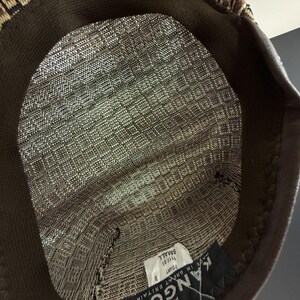 May include: The interior of a brown and tan patterned hat featuring a dark brown sweatband. A white tag is sewn inside with the text 'KANGOL', 'MADE IN GREAT BRITAIN', '2 DART', '1117.01', and 'SMALL' printed in black ink on the fabric label.