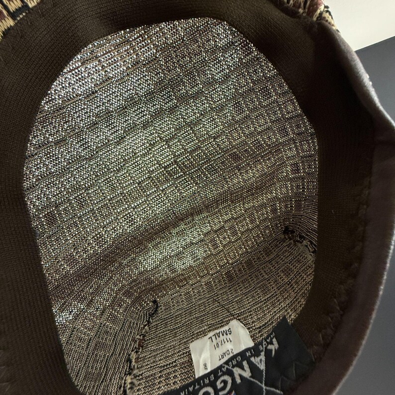 May include: The interior of a brown and tan patterned hat featuring a dark brown sweatband. A white tag is sewn inside with the text 'KANGOL', 'MADE IN GREAT BRITAIN', '2 DART', '1117.01', and 'SMALL' printed in black ink on the fabric label.
