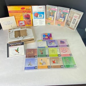 以下が含まれることがあります： 「creative fantasy CAP」と書かれた箱や「PFAFF」のロゴが入ったCDなど、ミシンアクセサリーとソフトウェアのコレクション。ミシンアタッチメントと「Amazing Designs」というタイトルの本も含まれています。