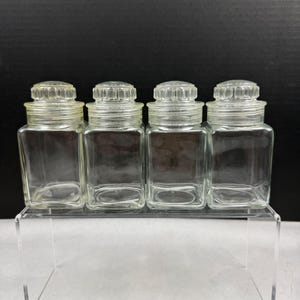 May include: Four clear glass spice jars with square bodies and ribbed lids. The jars are empty and sit on a clear acrylic shelf against a black background. The jars are vintage and appear to be in good condition.