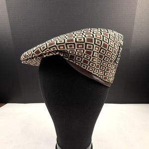 May include: A flat cap featuring a geometric diamond pattern in shades of brown, black, and cream. The cap has a structured crown and a dark brown leather-like trim along the bottom edge, with a small rectangular brand tag on the side.