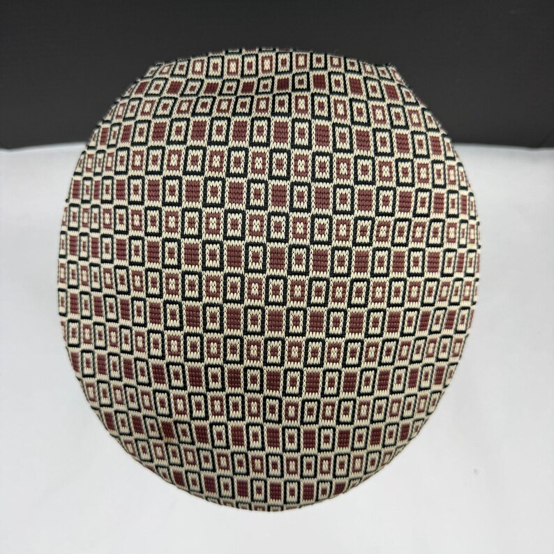 May include: A circular, flat-topped hat featuring a repeating geometric pattern of small, brown, black, and cream-coloured rectangles. The knit fabric displays a structured, grid-like design that covers the entire surface of the rounded headwear.