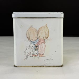 May include: A rectangular white metal tin with a lid. The tin features a vintage illustration of two children holding hands with a lamb. The children are wearing blue and pink clothing. The artist's signature, "Betsy Clark," is visible.