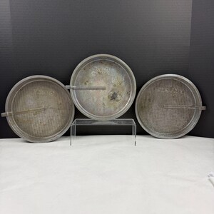 May include: Three round silver-colored metal cake pans with a textured grid pattern on the base. Each pan features the embossed text OVENEX and a small side handle. The pans are arranged with one elevated on a clear stand and two placed below.