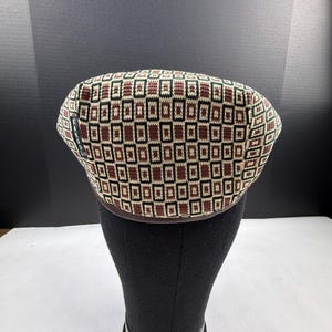 May include: A Kangol flat cap featuring a geometric pattern of brown, black, and cream-coloured rectangles. The knit fabric displays a repeating grid design across the crown, with a small black brand tag visible on the left side of the cap.