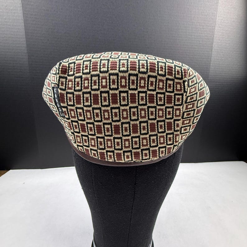 May include: A Kangol flat cap featuring a geometric pattern of brown, black, and cream-coloured rectangles. The knit fabric displays a repeating grid design across the crown, with a small black brand tag visible on the left side of the cap.
