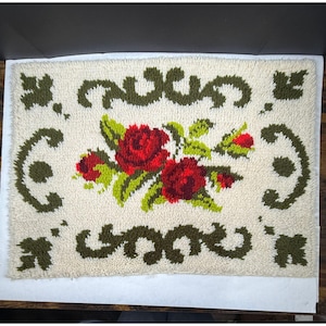May include: Rectangular off-white rug with a floral design. The rug features a central bouquet of red roses with green leaves. Dark green scrollwork borders the floral arrangement. The rug is likely made of a textured material.