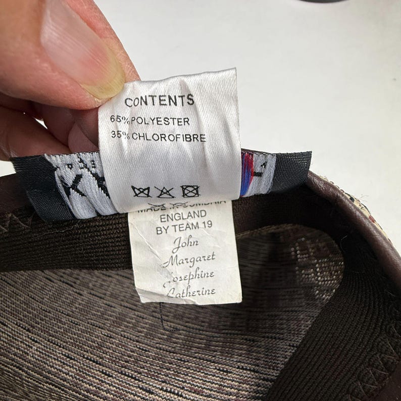 May include: A white fabric garment care label with black text reading CONTENTS 65% POLYESTER 35% CHLOROFIBRE, MADE IN ENGLAND BY TEAM 19, and the names John, Margaret, Josephine, and Catherine. The label includes three crossed-out laundry symbols.