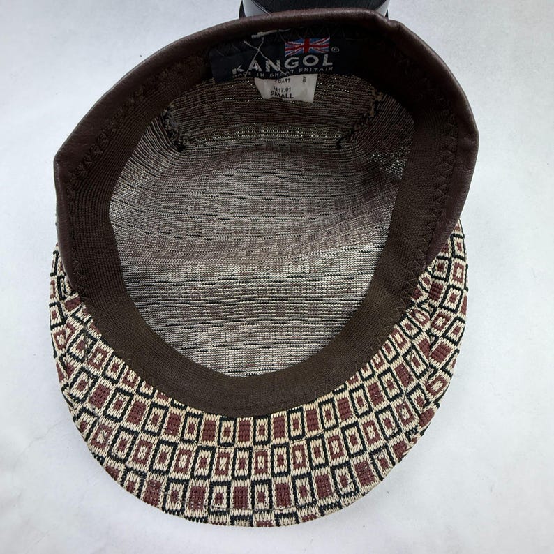 May include: A Kangol brand flat cap viewed from the interior, featuring a brown, black, and cream geometric square pattern. The inner band is dark brown, and a white label displays the text Kangol, Made in Great Britain, and Small.