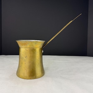 May include: A vintage brass coffee pot with a long, slender handle. The pot has a flared base and a narrow neck, with a silver-colored rim. The brass has a warm, golden hue, suggesting age and use. The handle extends upwards and outwards.