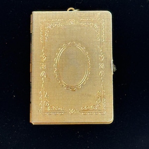 May include: A rectangular gold-toned metal compact case featuring a textured surface with an embossed decorative border and a central oval frame. The item includes a small metal loop at the top edge and a side latch mechanism for opening the case.