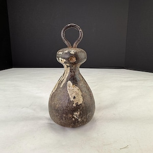 Puede incluir: Una pesa de hierro fundido vintage con un asa de bucle. La pesa marrón oscuro tiene salpicaduras de pintura blanca y una forma bulbosa. El asa es un bucle simple y redondeado. La pieza tiene una apariencia rústica y antigua.