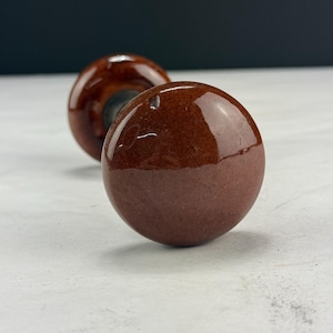 May include: Two brown, spherical cabinet knobs with a glossy finish. The knobs are a deep reddish-brown color, with a lighter, matte finish on the lower half. The knobs are likely made of ceramic or a similar material.