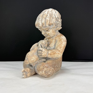 Puede incluir: Estatua beige de un niño sentado abrazando a un conejo. El niño mira hacia abajo al conejo, con una expresión serena. La estatua tiene un acabado texturizado, y los detalles de los rasgos del niño y el pelaje del conejo son visibles.