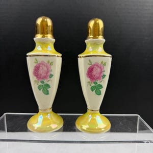 Pode incluir: Um par de saleiros e pimenteiros vintage. Os saleiros e pimenteiros são brancos sujos com um desenho de rosa rosa e detalhes dourados. Os topos e as bases são dourados. Os saleiros e pimenteiros têm cerca de 10 cm de altura.