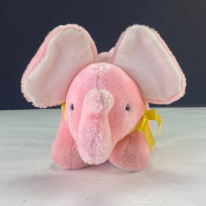 Puede incluir: Un juguete de elefante rosa de peluche con orejas grandes y una trompa. El elefante tiene ojos azules pequeños y una cinta amarilla alrededor del cuello. El juguete está hecho de material suave y difuso.