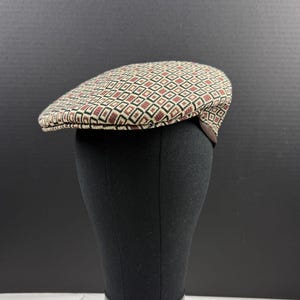 May include: A flat cap featuring a repeating geometric pattern of small squares in shades of tan, dark brown, and black. The hat has a structured, rounded crown and a short, stiff brim that extends from the front, displaying a woven texture.