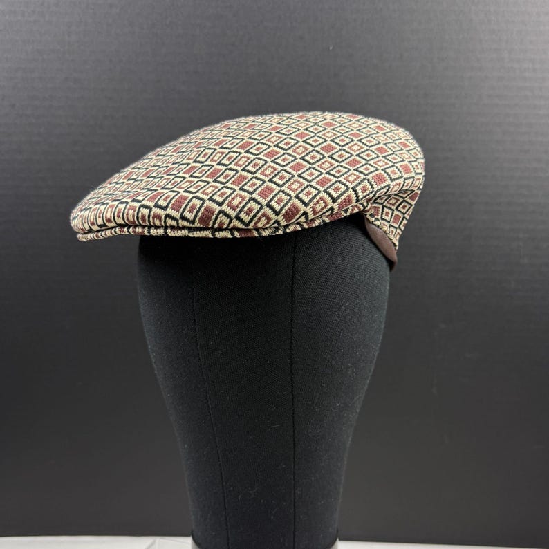May include: A flat cap featuring a repeating geometric pattern of small squares in shades of tan, dark brown, and black. The hat has a structured, rounded crown and a short, stiff brim that extends from the front, displaying a woven texture.