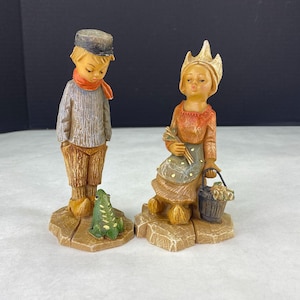 Puede incluir: Dos figuras de cerámica. Uno es un niño con sombrero, bufanda y pantalón marrón, con una rana verde a sus pies. La otra es una niña con corona, que sostiene una cesta y un manojo de palos. Ambos están sobre bases texturizadas.