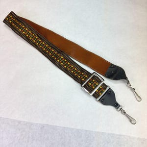 May include: A camera strap featuring a woven geometric pattern in shades of orange, yellow, and green. The strap includes a brown leather underside, a silver-toned metal adjustment buckle, and black leather end tabs with silver-toned metal swivel clips.