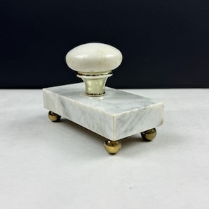 May include: A rectangular marble ink blotter with a white and gray pattern. It has a rounded, off-white handle and sits on four small, gold-colored ball feet. The handle is attached to a gold-colored base.