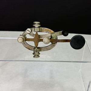 May include: A vintage telegraph key featuring an oval-shaped metal frame with a weathered patina. The device includes multiple adjustable knurled knobs, a central cross-shaped component, and a prominent black circular knob extending to the right side.