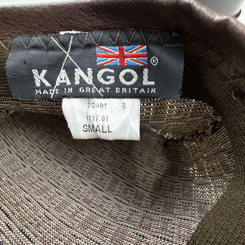 May include: A brown Kangol hat with a dark brown leather trim. The interior features a black fabric label with the text KANGOL, MADE IN GREAT BRITAIN, and a Union Jack flag. A white tag below displays the text 2 DART, 1117.01, and SMALL.