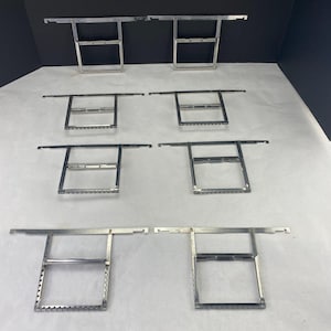 Puede incluir: Colección de siete escuadras de estantería de metal plateado. Cada escuadra tiene un marco rectangular con una barra horizontal que se extiende a ambos lados. Las escuadras están dispuestas sobre una superficie blanca.