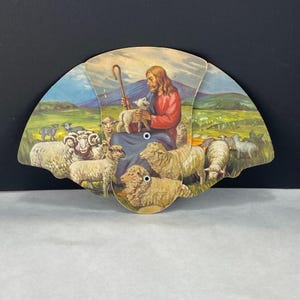 May include: A vintage fan depicting a religious scene with a figure in red and blue robes holding a lamb, surrounded by sheep. The background shows a green landscape with mountains and a blue sky. The fan is held together with two small rivets.