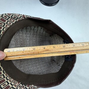 May include: A flat cap with a brown, black, and cream geometric pattern. The interior features a dark brown sweatband and a black fabric label. A wooden ruler is placed across the opening, showing a measurement of approximately 20.3 cm.