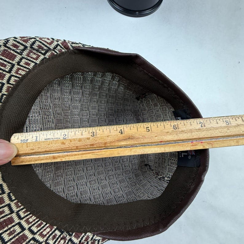 May include: A flat cap with a brown, black, and cream geometric pattern. The interior features a dark brown sweatband and a black fabric label. A wooden ruler is placed across the opening, showing a measurement of approximately 20.3 cm.